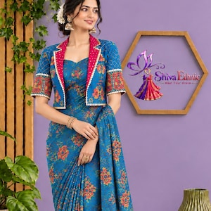 Op de afbeelding: Een blauwe sari met een herhalend oranje en rood bloemenpatroon, gecombineerd met een bijpassend jasje met korte mouwen. Het jasje heeft een felroze revers met witte stippen en gouden randen. De tekst Shiva Ethnic Wear Your Grace is zichtbaar.
