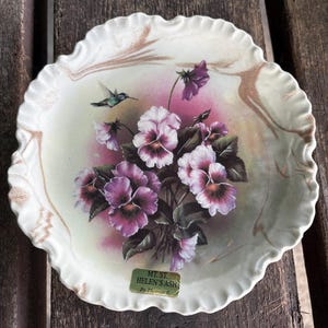 May include: A round, scalloped-edge decorative plate featuring a central illustration of purple and white pansies with green leaves and a small hummingbird. The plate has a cream-colored background with gold accents and a label reading MT. ST. HELEN'S ASH By Thomas E.