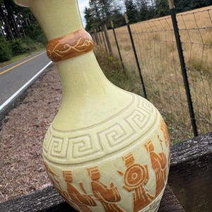 May include: A pale yellow ceramic vase with a narrow neck and a bulbous body. The vase features a raised brown relief design depicting figures in traditional attire and a geometric Greek key pattern around the neck and shoulder areas.
