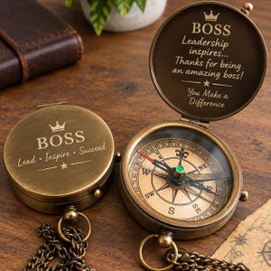 Puede incluir: Dos brújulas de latón de estilo antiguo con cadenas. Una está cerrada, grabada con una corona, 'BOSS' y 'Lead • Inspire • Succeed'. La otra está abierta, revelando un dial direccional y una tapa interior grabada con 'BOSS', 'Leadership inspires...', 'Thanks for being an amazing boss!' y 'You Make a Difference'.