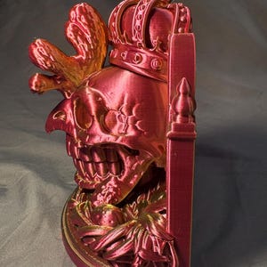 Side view of King of the Coop in red and gold tones, showing its rooster crest, crowned skull, shallow-backed form and flat display base. This is an indoor decorative sculpture.