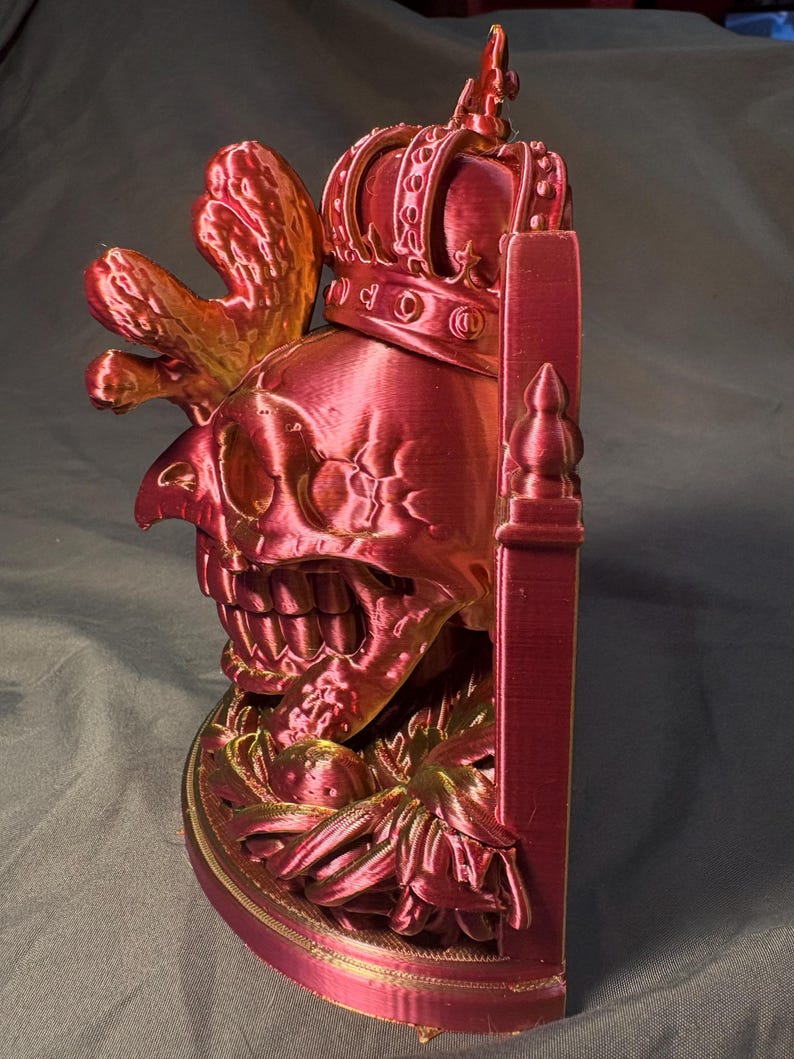 Side view of King of the Coop in red and gold tones, showing its rooster crest, crowned skull, shallow-backed form and flat display base. This is an indoor decorative sculpture.
