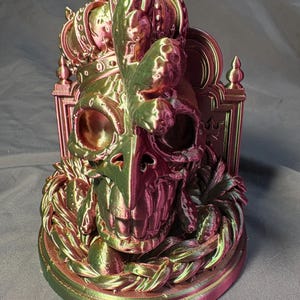 Front-left view of the same King of the Coop sculpture. The crowned rooster-inspired skull shows green, gold and magenta tones in the silk PLA, with visible print layers and a rounded display base.