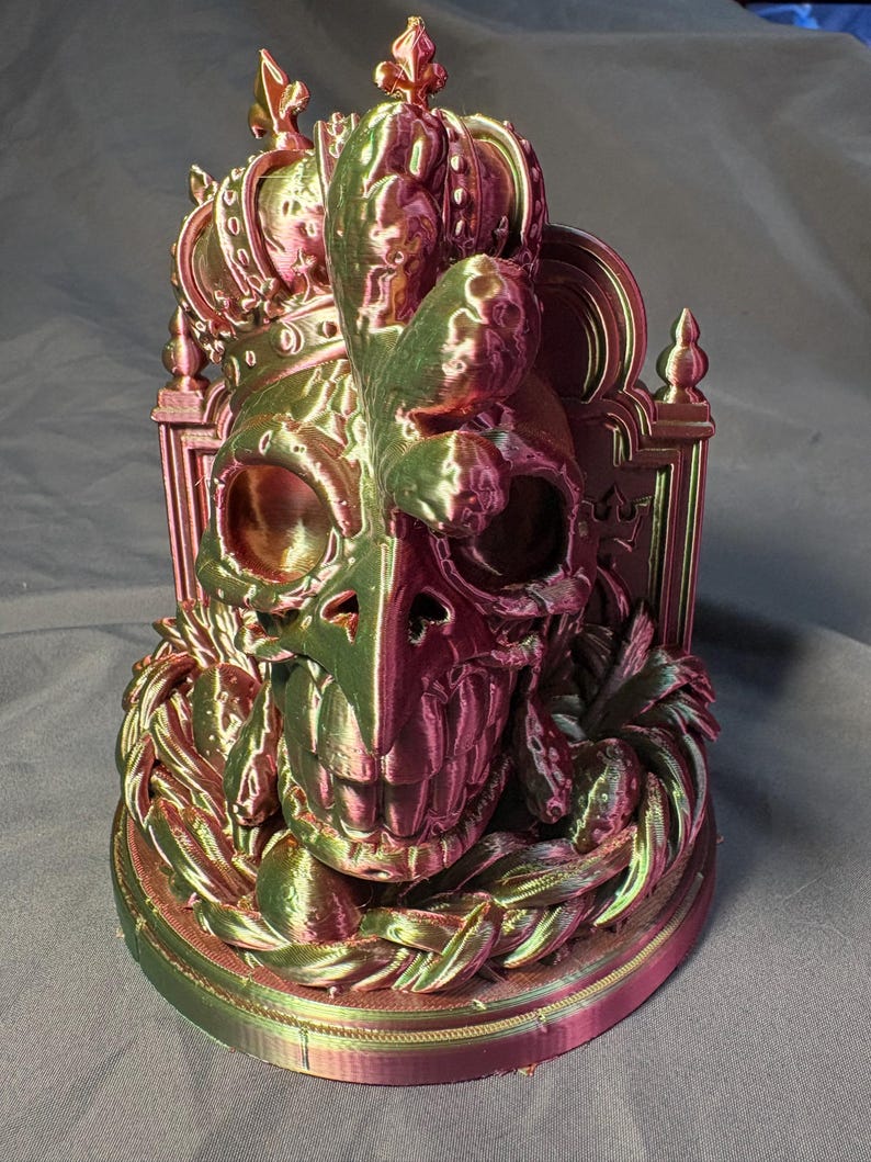 Front-left view of the same King of the Coop sculpture. The crowned rooster-inspired skull shows green, gold and magenta tones in the silk PLA, with visible print layers and a rounded display base.