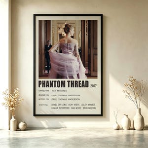 May include: A framed movie poster for the 2017 film Phantom Thread. The poster features a central image of a person in a light purple gown standing in a doorway, with black text listing the title, year, director, writer, and cast members.