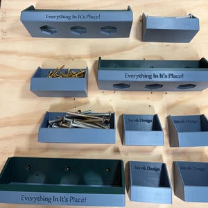 May include: A collection of grey and dark green storage bins of varying sizes, some containing metal screws. Several bins feature the text "Everything In It's Place!" or "Stirvik Design" embossed on the front. The bins have open tops and angled sides.