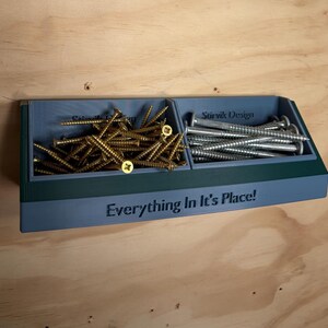 May include: A blue, wall-mounted storage bin with two compartments, featuring the text "Everything In It's Place!" on the front and "Stirvik Design" inside. The left compartment holds gold-coloured screws, while the right holds silver-coloured screws.