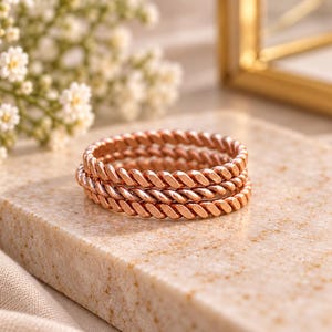 Op de afbeelding: Een stapel van drie roségouden ringen, elk met een doorlopende gedraaide touwtextuur. De ringen zijn uniform in grootte en ontwerp en vertonen een metallic koperkleurige afwerking met een herhalend diagonaal patroon langs de gehele omtrek.