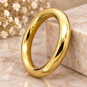 May include: A polished gold-tone ring with a smooth, rounded, and thick band. The ring features a continuous, seamless circular design with a highly reflective metallic surface that catches the light, creating bright highlights along its curved edges.