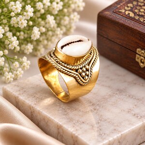 Puede incluir: Un anillo de tono dorado que presenta una concha de cauri blanca central engastada en un marco de metal decorativo. La banda está elaborada con una textura de cuerda trenzada y pequeños acentos de cuentas esféricas alrededor de la concha.