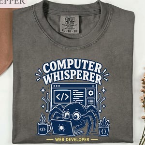 May include: A charcoal gray t-shirt featuring a graphic of a spider wearing glasses while typing on a laptop. The text reads Computer Whisperer and Web Developer. The design includes a coffee mug, a potted plant, and code symbols.