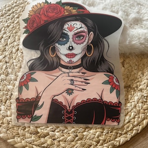 Könnte beinhalten: Ein Aufkleber mit einer stilisierten Illustration einer Person mit Sugar-Skull-Gesichtsbemalung, die einen breitkrempigen schwarzen Hut mit roten Rosen und Sonnenblumen trägt. Die Figur hat langes dunkles Haar, goldene Creolen, ein schwarzes Halsband und Tattoos.