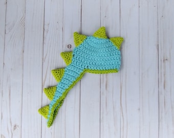 May include: A crocheted dinosaur hat with a blue and green tail. The hat is made of yarn and has a pointed top.