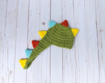 May include: Green crocheted dinosaur hat with a long tail and colourful spikes. The hat is made of yarn and has a pointed top.