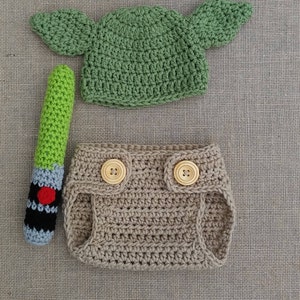 Może przedstawiać: Zielony, szydełkowy kapelusz Yody z opadającymi uszami, brązowy, szydełkowy pokrowiec na pieluchę z drewnianymi guzikami i zielono-czarna, szydełkowa zabawka w kształcie miecza świetlnego.