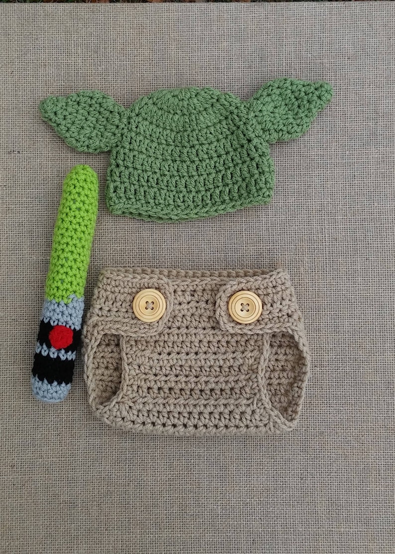 Może przedstawiać: Zielony, szydełkowy kapelusz Yody z opadającymi uszami, brązowy, szydełkowy pokrowiec na pieluchę z drewnianymi guzikami i zielono-czarna, szydełkowa zabawka w kształcie miecza świetlnego.