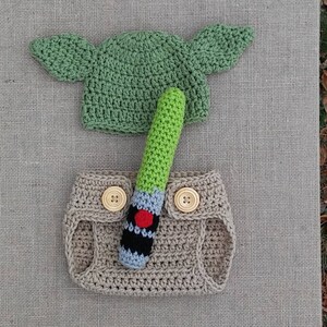 Może przedstawiać: Ręcznie robiony str&oacute;j dla niemowlęcia inspirowany Gwiezdnymi Wojnami, z zielonym kapeluszem Yody, brązową pieluchą i zielonym mieczem świetlnym.