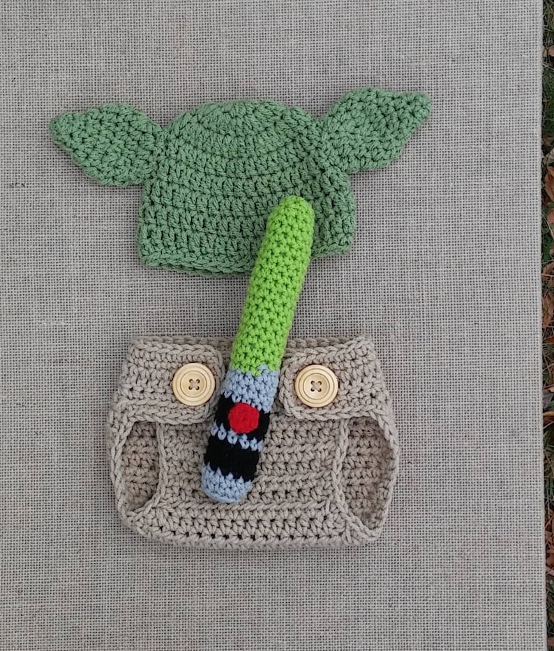 Może przedstawiać: Ręcznie robiony str&oacute;j dla niemowlęcia inspirowany Gwiezdnymi Wojnami, z zielonym kapeluszem Yody, brązową pieluchą i zielonym mieczem świetlnym.