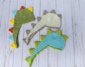 May include: Three crocheted dinosaur hats in green, beige, and blue with yellow, orange, and green accents. The hats have long tails that resemble dinosaur tails.