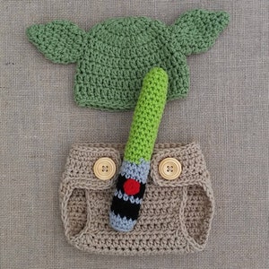 Może przedstawiać: Zielony, dziany kapelusz z uszami, brązowy, dziany pieluch i zielono-czarna, dziana szabla świetlna.