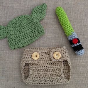 Może przedstawiać: Zielony, dziany kapelusz z ostrymi uszami, brązowe, dziane pieluchy z dwoma drewnianymi guzikami i zielona, szara i czarna, dziana szabla świetlna.