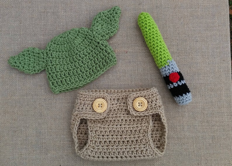 Może przedstawiać: Zielony, dziany kapelusz z ostrymi uszami, brązowe, dziane pieluchy z dwoma drewnianymi guzikami i zielona, szara i czarna, dziana szabla świetlna.