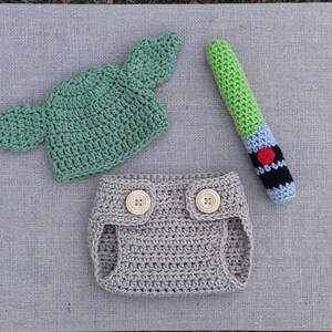 Może przedstawiać: Ubranko dla niemowlęcia wykonane na szydełku z zielonym kapeluszem Yody, brązową osłoną na pieluchę i zieloną zabawką w kształcie miecza świetlnego.