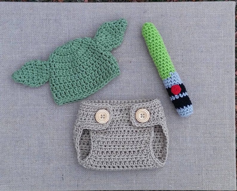 Może przedstawiać: Ubranko dla niemowlęcia wykonane na szydełku z zielonym kapeluszem Yody, brązową osłoną na pieluchę i zieloną zabawką w kształcie miecza świetlnego.