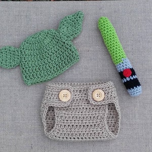 Może przedstawiać: Zielony, szydełkowy kapelusz Yody z dużymi uszami, beżowa, szydełkowa osłonka na pieluchę z dwoma drewnianymi guzikami i zielono-niebieski, szydełkowy miecz świetlny.