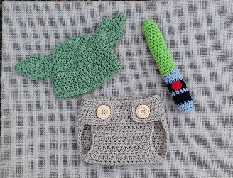 Może przedstawiać: Zielony, szydełkowy kapelusz Yody z dużymi uszami, beżowa, szydełkowa osłonka na pieluchę z dwoma drewnianymi guzikami i zielono-niebieski, szydełkowy miecz świetlny.