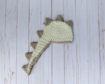 May include: A crocheted dinosaur hat with a long tail. The hat is beige and brown with a textured stitch.