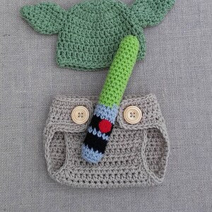 Może przedstawiać: Ubranko dla niemowlęcia wykonane na szydełku w kolorze zielonym, brązowym i niebieskim. Ubranko zawiera czapkę z uszami, osłonę na pieluchę i zabawkę w kształcie miecza świetlnego.