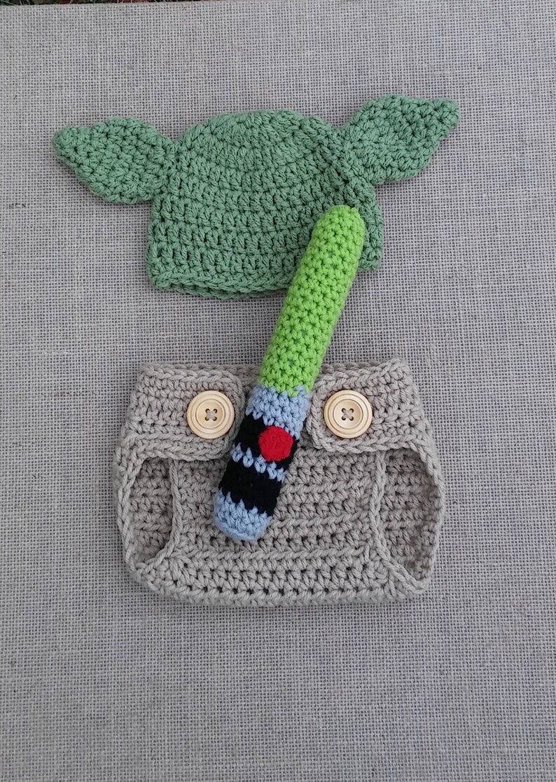 Może przedstawiać: Ubranko dla niemowlęcia wykonane na szydełku w kolorze zielonym, brązowym i niebieskim. Ubranko zawiera czapkę z uszami, osłonę na pieluchę i zabawkę w kształcie miecza świetlnego.