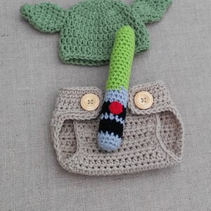 Może przedstawiać: Kostium dla niemowlęcia wykonane na szydełku w kolorze beżowym z dwoma brązowymi guzikami i zieloną czapką na szydełku z ostrymi uszami. W zestawie znajduje się zielony, czarny i szary miecz świetlny wykonany na szydełku.