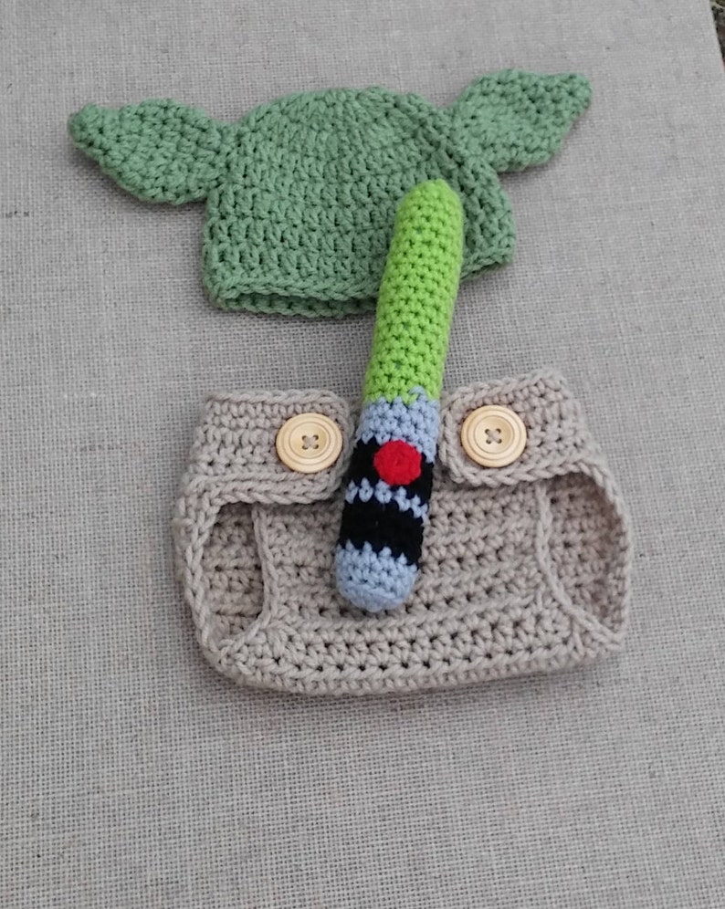 Może przedstawiać: Kostium dla niemowlęcia wykonane na szydełku w kolorze beżowym z dwoma brązowymi guzikami i zieloną czapką na szydełku z ostrymi uszami. W zestawie znajduje się zielony, czarny i szary miecz świetlny wykonany na szydełku.