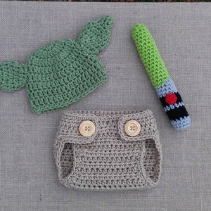 Może przedstawiać: Kr&oacute;liczy str&oacute;j dla niemowlęcia w kolorze zielonym i beżowym, w skład kt&oacute;rego wchodzi czapka z uszami, osłona na pieluchę i zabawka w kształcie miecza świetlnego.