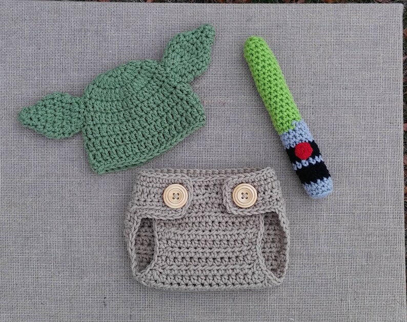 Może przedstawiać: Kr&oacute;liczy str&oacute;j dla niemowlęcia w kolorze zielonym i beżowym, w skład kt&oacute;rego wchodzi czapka z uszami, osłona na pieluchę i zabawka w kształcie miecza świetlnego.