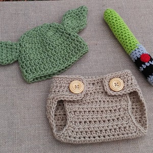 Może przedstawiać: Ubranko dla niemowlęcia wykonane na szydełku z zielonym kapeluszem, brązowymi pieluchami i zieloną zabawką w kształcie miecza świetlnego.