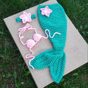 May include: A crocheted mermaid tail costume set in teal and pink. The set includes a tail, a top, and a headband. The headband and top are decorated with pink flowers.