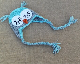 May include: A blue and gray crocheted owl hat with a long braided cord. The owl has white eyes and an orange beak.