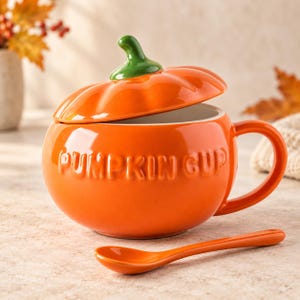 Könnte beinhalten: Ein leuchtend orangefarbener Keramikbecher in Kürbisform mit passendem Deckel und grünem Stiel. Das Wort PUMPKIN CUP ist auf der Vorderseite eingeprägt. Ein passender orangefarbener Löffel liegt vor dem Becher, der einen geschwungenen Henkel hat.