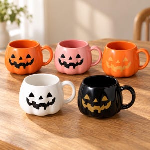 Könnte beinhalten: Ein Set aus fünf kürbisförmigen Keramiktassen, die jeweils mit einem einzigartigen Jack-o'-Lantern-Gesicht verziert sind. Das Set besteht aus einer leuchtend orangen, einer pastellrosa, einer orangen mit hellem Gesicht, einer weißen und einer schwarzen Tasse.