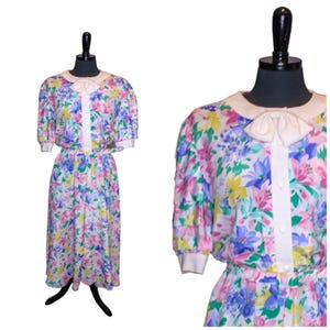 May include: A white dress with a floral print in shades of pink, yellow, blue, and green. The dress has a white collar and a bow at the neck. The dress has short sleeves and a button-up front.