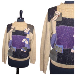 Puede incluir: Un suéter beige con cuello alto y patchwork. El suéter presenta cuadrados de tela morada, gris y azul marino. Hojas y bayas aplicadas adornan la parte delantera. El suéter se exhibe en un maniquí negro.