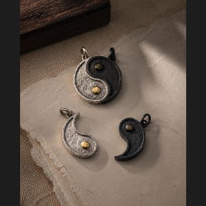 May include: Three metal pendants featuring a yin-yang design. One circular pendant shows a split black and silver textured surface with small gold-toned accents. Two separate comma-shaped pendants, one silver and one black, each feature a gold-toned dot.