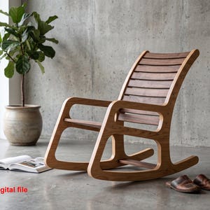 Puede incluir: Una mecedora de madera con respaldo y asiento de listones, con un marco de tono claro y listones de madera más oscura. La silla tiene balancines y reposabrazos curvos. El texto "digital file" está impreso en rojo en la esquina inferior izquierda.