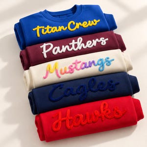 Può includere: Una pila di cinque felpe a girocollo piegate nei colori blu, bordeaux, crema, blu navy e rosso. Ogni felpa presenta sul davanti un ricamo in rilievo e testurizzato con le parole Titan Crew, Panthers, Mustangs, Cagles e Hawks in colori contrastanti.