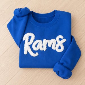 May include: A royal blue crewneck sweatshirt featuring the word Rams in large, white, textured chenille-style script lettering across the chest. The garment has long sleeves with ribbed cuffs and a ribbed neckline, providing a classic pullover design.