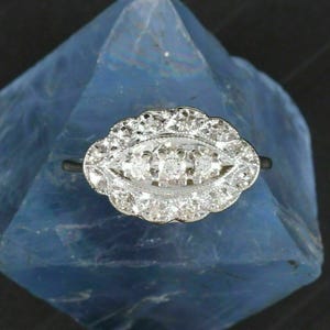 May include: A silver-toned ring featuring an elongated, scalloped oval setting. The centre of the ring contains three small, round, clear stones arranged horizontally, surrounded by a border of numerous smaller, sparkling clear stones.