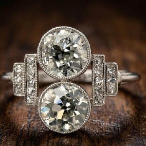 Puede incluir: Un anillo de tono plateado con dos piedras redondas transparentes apiladas verticalmente. El engaste presenta piedras redondas más pequeñas incrustadas en paneles laterales rectangulares, todo con un delicado detalle de borde milgrain.